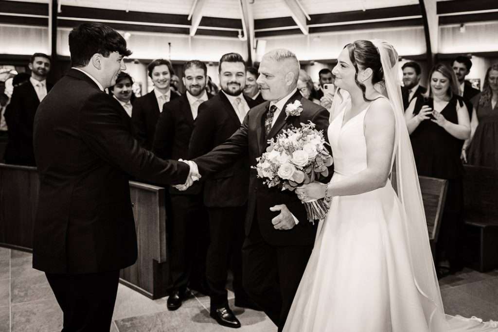 Bride’s father shakes hands with the groom at the altar during a heartfelt moment, with the bride holding her bouquet and smiling beside them.