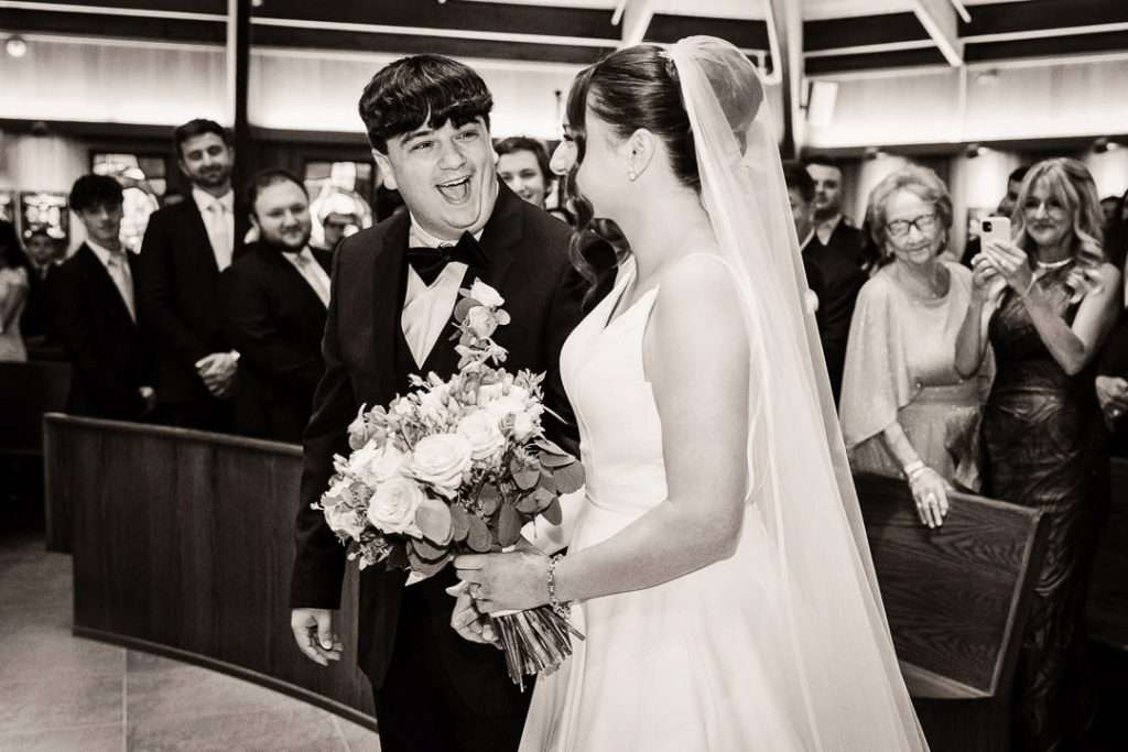 Groom beams with joy as he sees his bride at the altar, surrounded by smiling guests and groomsmen at Notre Dame of Mt. Carmel.