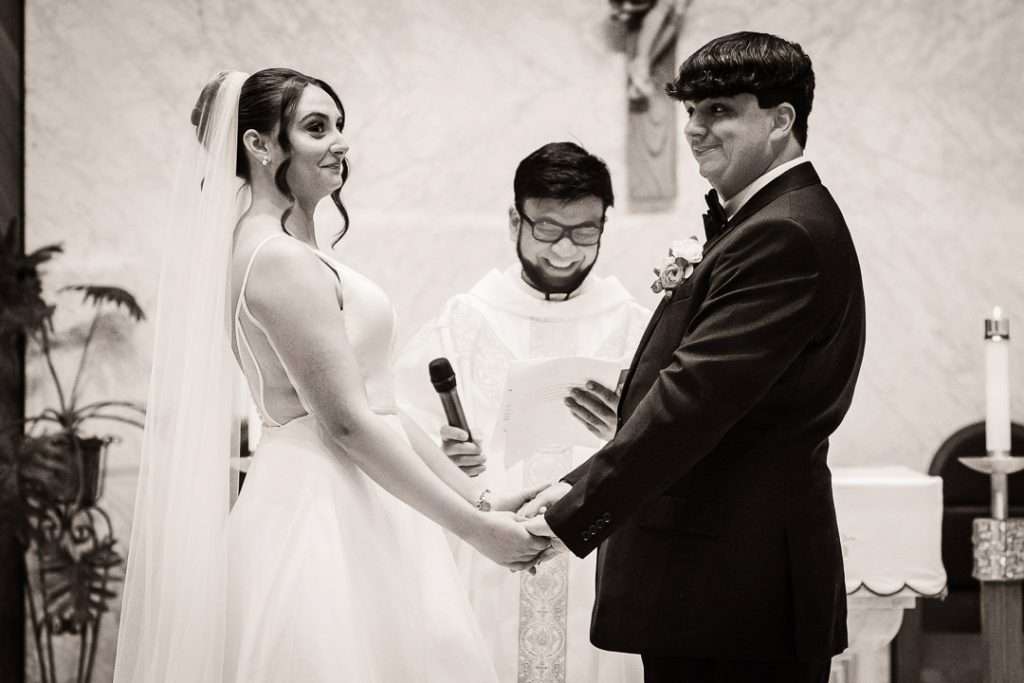 Bride and groom hold hands and share a smile at the start of their wedding ceremony as the officiant reads from his notes.