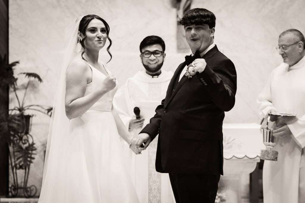 Bride and groom celebrate playfully holding hands in front of the altar, with joyful reactions and laughter from their priest and guests.
