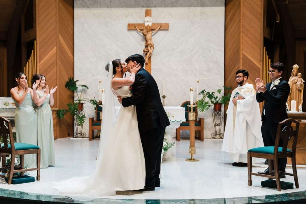 Bride and groom share their first kiss at the altar under the crucifix, surrounded by clapping bridal party and priest.
