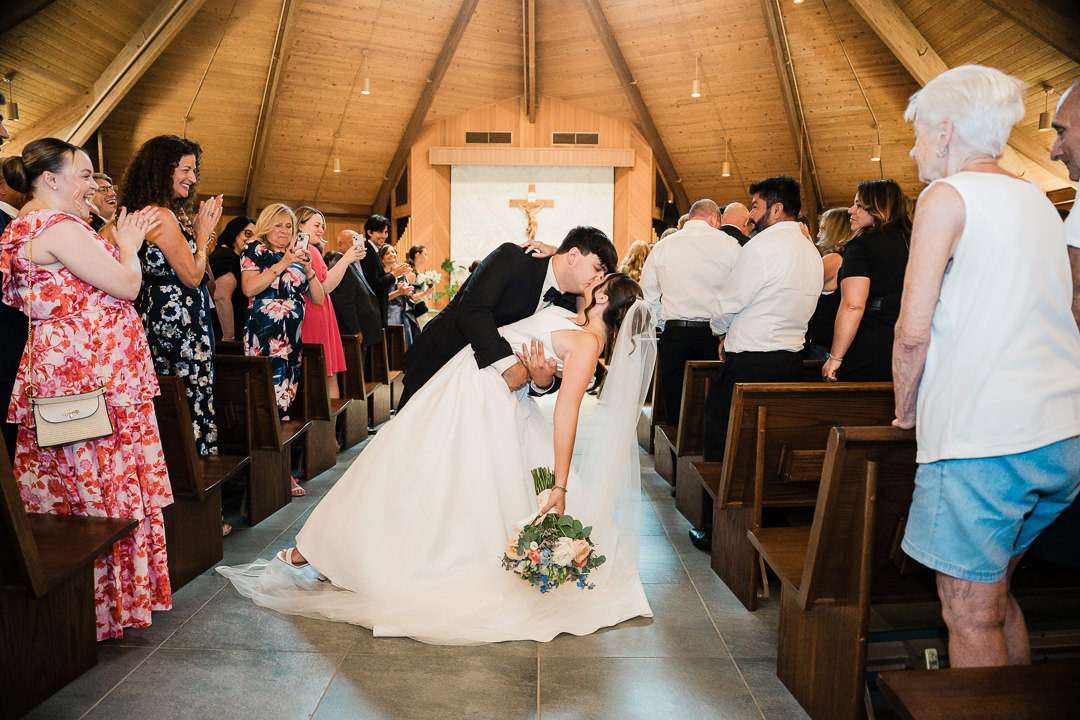 Bride and groom share a dramatic dip kiss while walking down the aisle at Notre Dame of Mt. Carmel Roman Catholic Church, surrounded by cheering guests in vibrant summer attire.