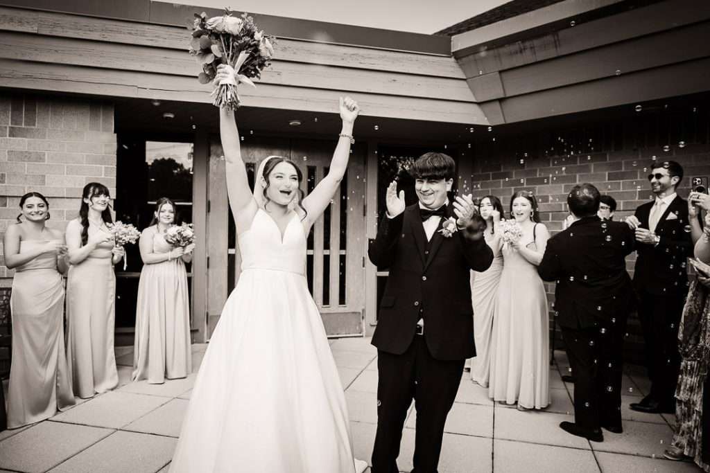 Bride cheers with arms raised during the bubble exit while the groom claps beside her, surrounded by the bridal party and guests.