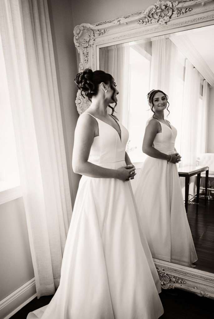 Bride gazes at her reflection in a full-length mirror, wearing a classic white gown in a bright bridal suite at The Mansion at Mountain Lakes.
