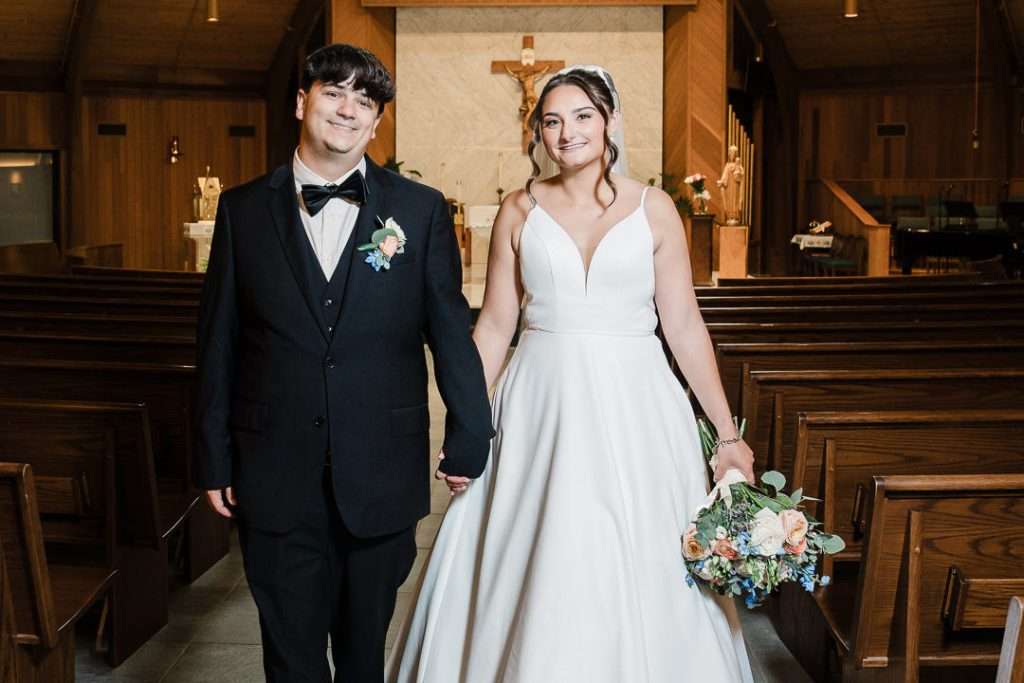 Bride and groom pose for a front-facing portrait while walking down the aisle, smiling in front of the altar at Notre Dame of Mt. Carmel.