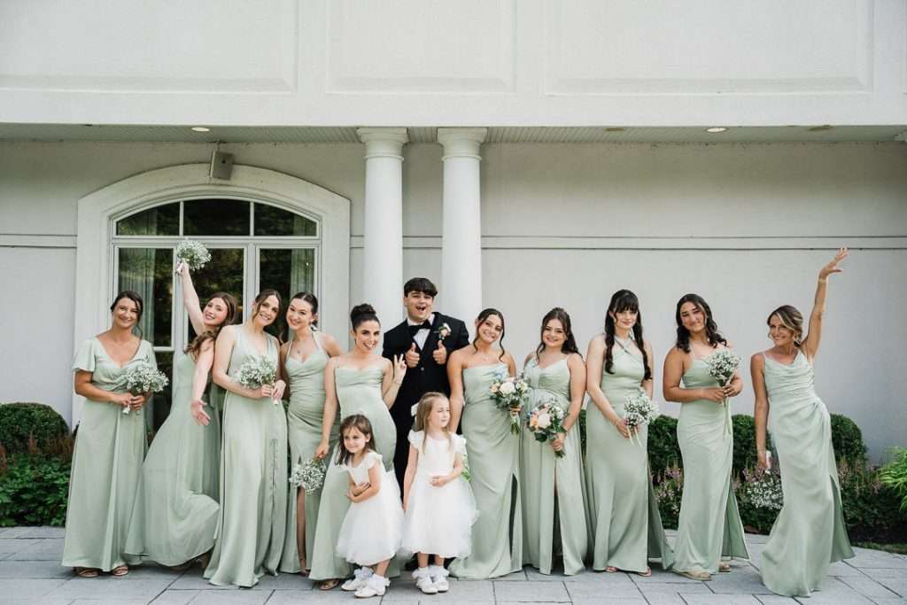 Groom kneels surrounded by laughing bridesmaids in matching dresses, all striking playful poses during a fun pre-ceremony moment.