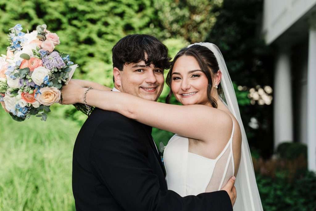 Bride and groom embrace outdoors, smiling at each other with soft natural light and greenery in the background at The Mansion at Mountain Lakes.