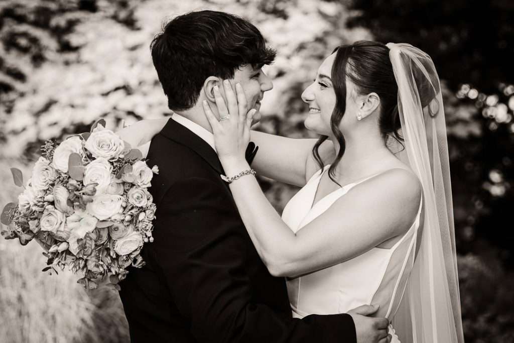 Bride gently holds groom’s face in a romantic close-up as they smile at each other during golden hour on the venue’s manicured lawn.