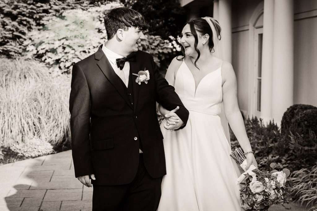 Bride and groom walk hand-in-hand, smiling at each other along a tree-lined path at The Mansion at Mountain Lakes.