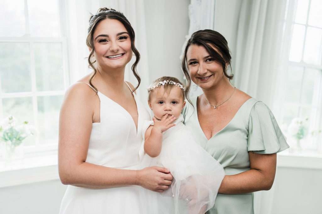 Bride poses with her mother and baby girl in a soft, light-filled room, all smiling in coordinated pastel outfits at The Mansion at Mountain Lakes.