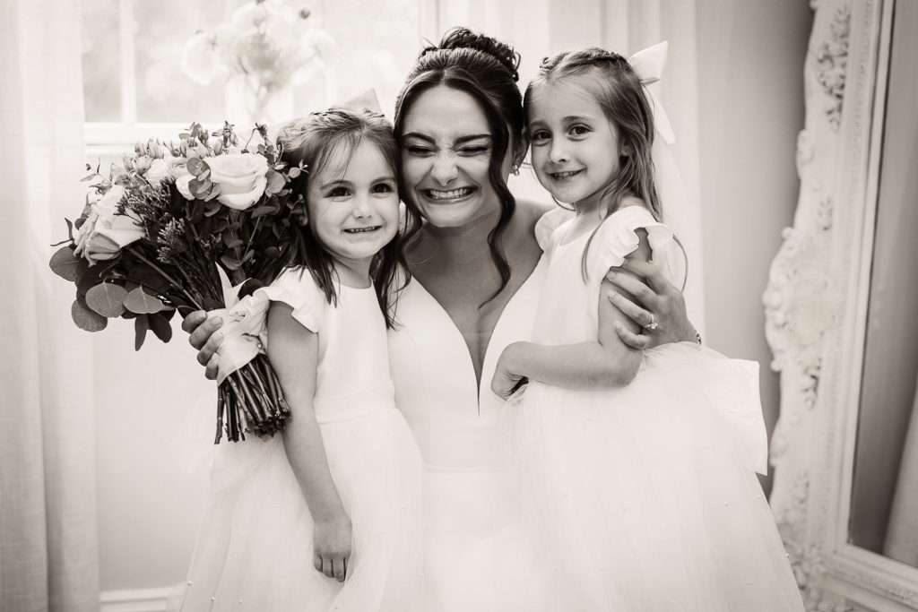 Emotional black-and-white photo of the bride hugging two smiling flower girls tightly, all in white dresses, bouquet in hand at The Mansion at Mountain Lakes.