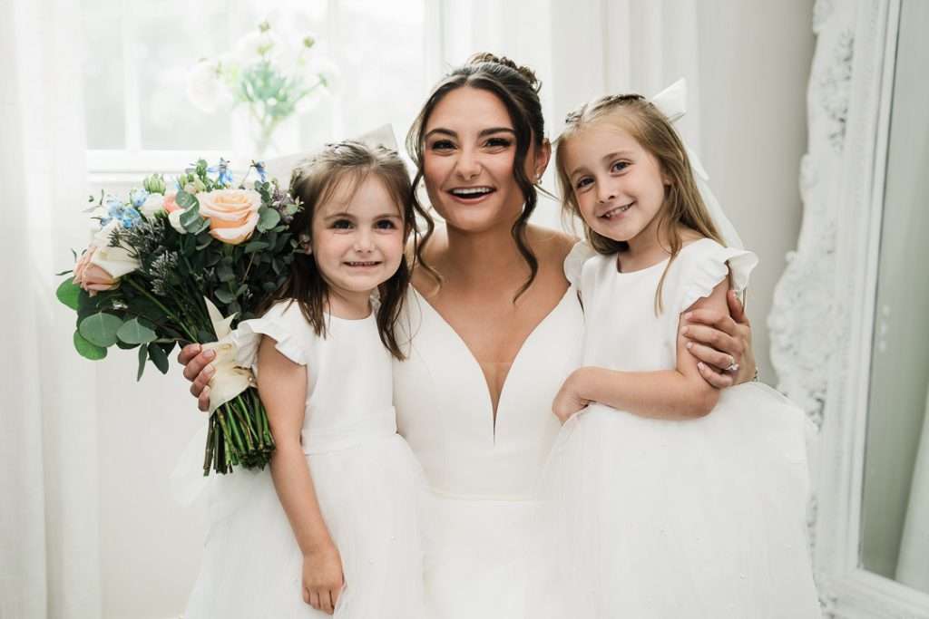 Bride poses happily between two flower girls in white dresses, holding a lush bouquet of blush, peach, and blue flowers at The Mansion at Mountain Lakes.