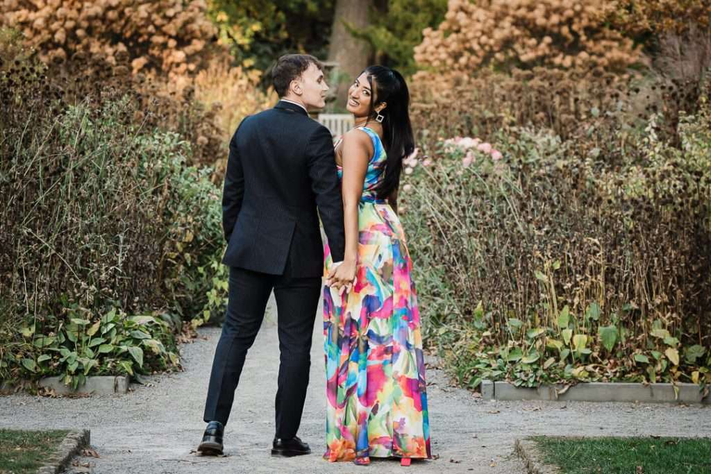 Engaged couple walking hand-in-hand through the garden at Van Vleck House, smiling back toward the camera. The woman wears a vibrant floral dress, adding a pop of color against the fall foliage backdrop.
