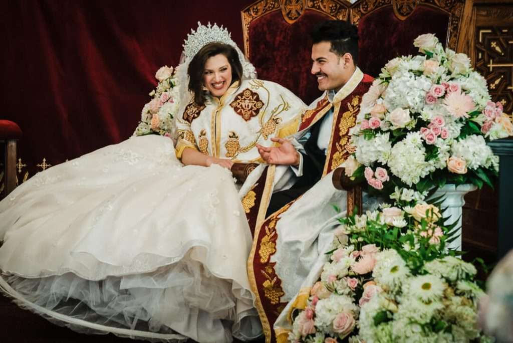 Bride and groom smiling while seated on thrones during a traditional Coptic wedding ceremony surrounded by lush floral arrangements.

