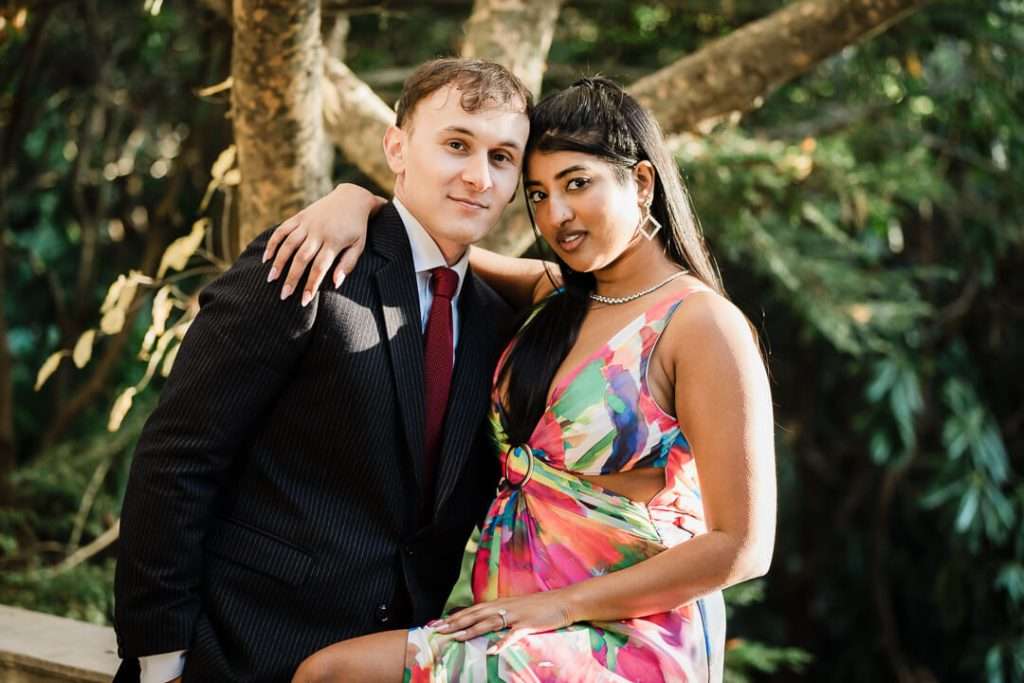 Engaged couple posing confidently at Van Vleck Gardens, with the woman in a vibrant multicolor dress and the man in a dark suit, framed by warm golden light and trees in the background.