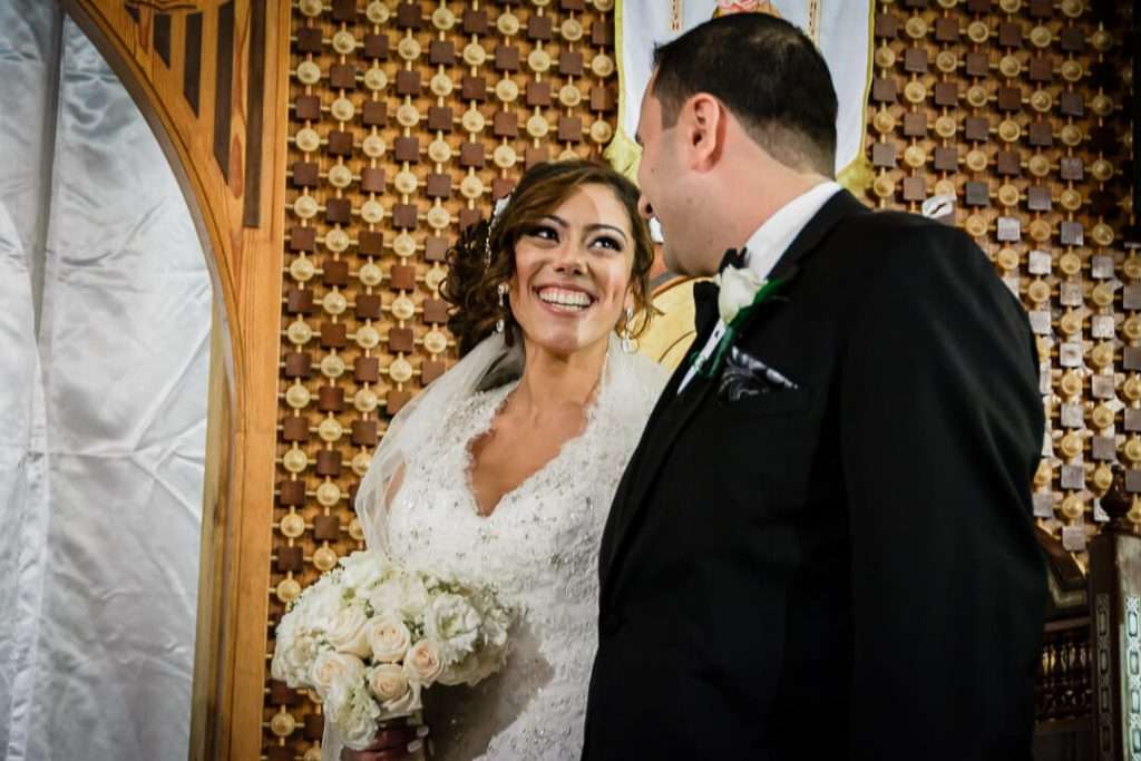 Coptic bride beaming at her groom while holding a bouquet of white roses during their wedding ceremony in front of a decorative altar.