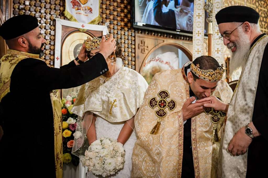 Coptic priest places a golden crown on the bride’s head while the groom kisses the priest’s hand in reverence during the traditional crowning ceremony.