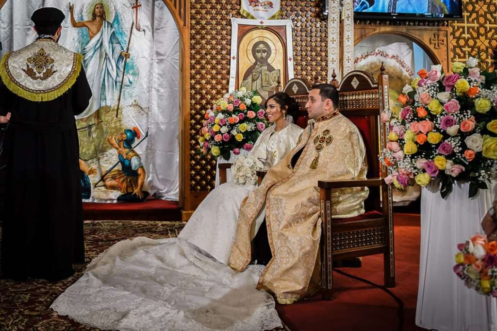 Bride and groom seated at the altar in full Coptic ceremonial attire as the priest leads the wedding ceremony, surrounded by colorful flower arrangements.