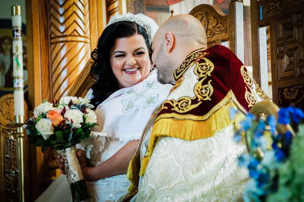 Bride smiles at groom while seated during Coptic Orthodox wedding, holding bouquet.
