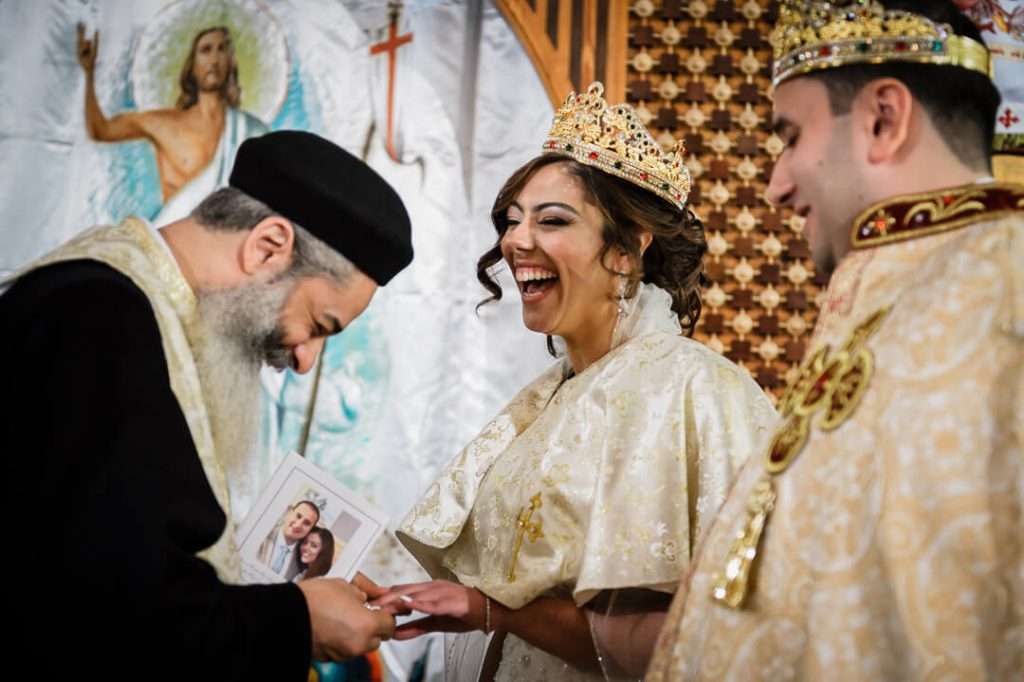 Coptic Orthodox priest places a ring on the bride’s finger as she laughs joyfully during the wedding ceremony, with the groom smiling beside her.