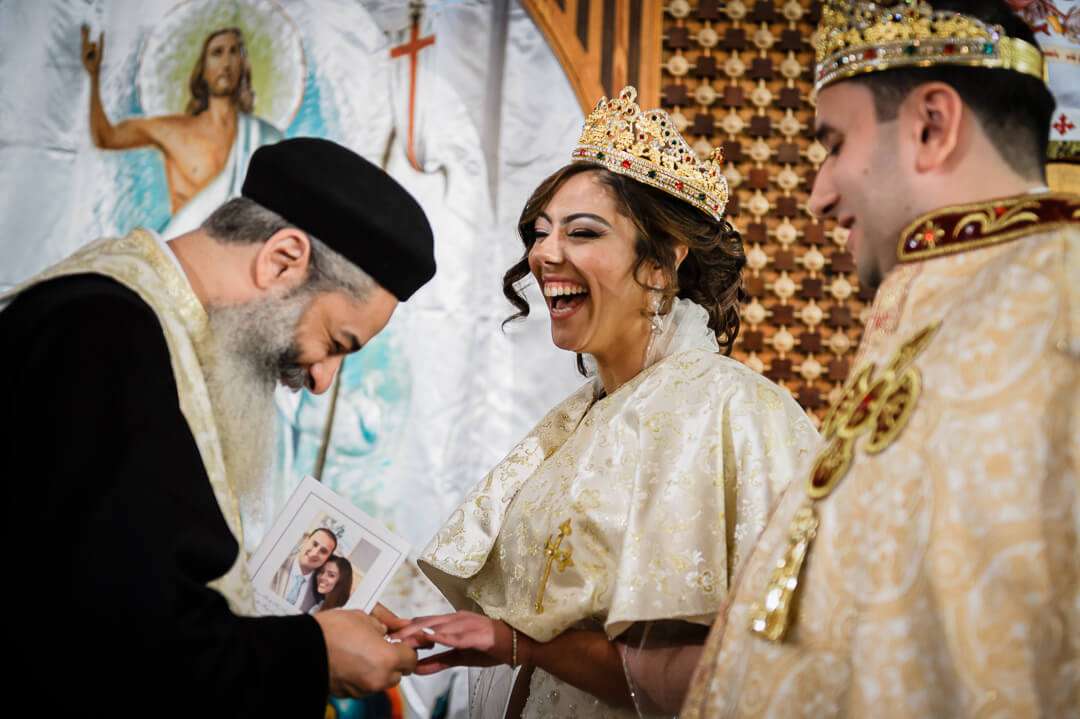 Coptic Orthodox priest places a ring on the bride’s finger as she laughs joyfully during the wedding ceremony, with the groom smiling beside her.
