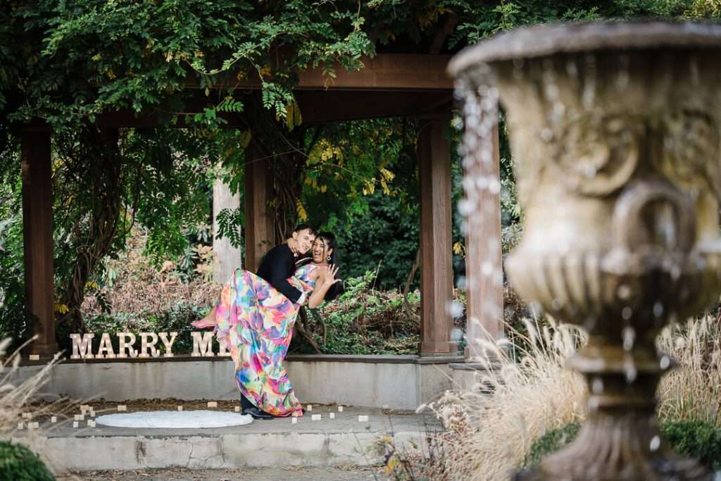Groom dips his fiancée for a kiss under the pergola at Van Vleck Gardens, framed by a classic stone fountain in the foreground.