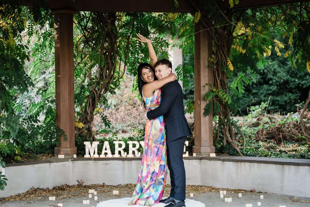 Engaged couple celebrating under the pergola at Van Vleck House with a “Marry Me” sign and candles, her hand raised in joy.
