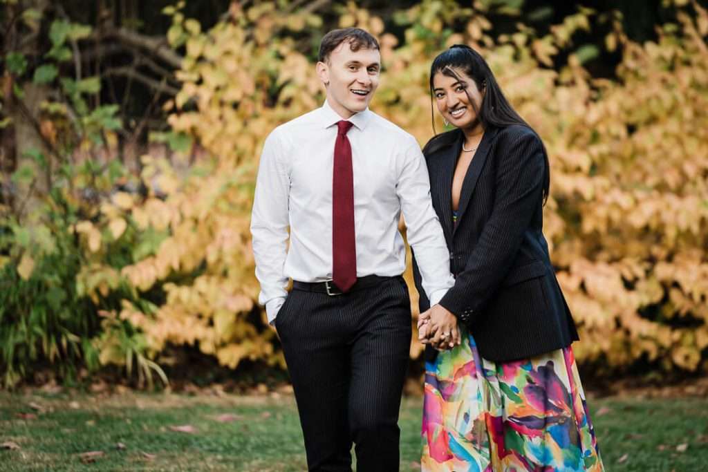 Engaged couple laughing together in front of golden autumn leaves at Van Vleck Gardens. The woman wears a colorful dress paired with a black blazer, and the man is in a white shirt with a red tie, both looking relaxed and joyful.
