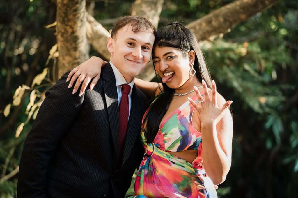 Playful moment at Van Vleck Gardens as newly engaged woman proudly shows off her engagement ring, smiling with her tongue out while leaning into her fiancé in a suit.