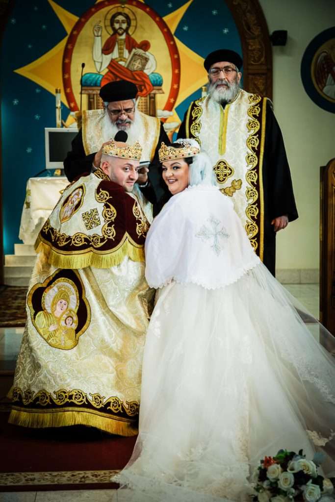 Smiling bride and groom crowned and kneeling during joyful Coptic Orthodox wedding in NJ