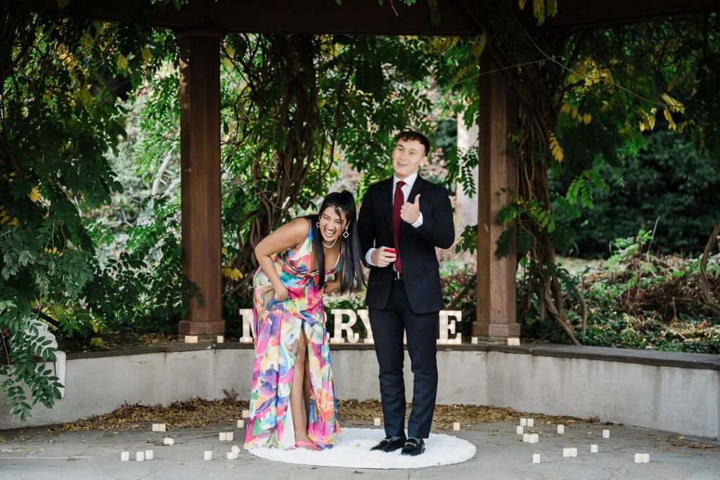 Couple laughing after a successful proposal at Van Vleck House, with the groom holding the red ring box and giving a thumbs up.