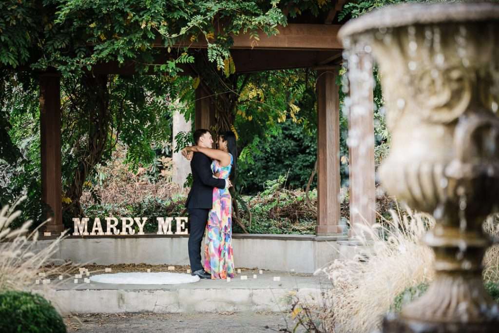 Engaged couple embracing under the pergola at Van Vleck House with “Marry Me” sign in the background and a soft-focus fountain in the foreground.