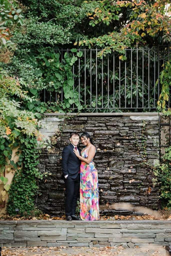 Engaged couple standing in front of a lush stone wall at Van Vleck Gardens, surrounded by greenery and autumn leaves, smiling warmly with arms around each other.