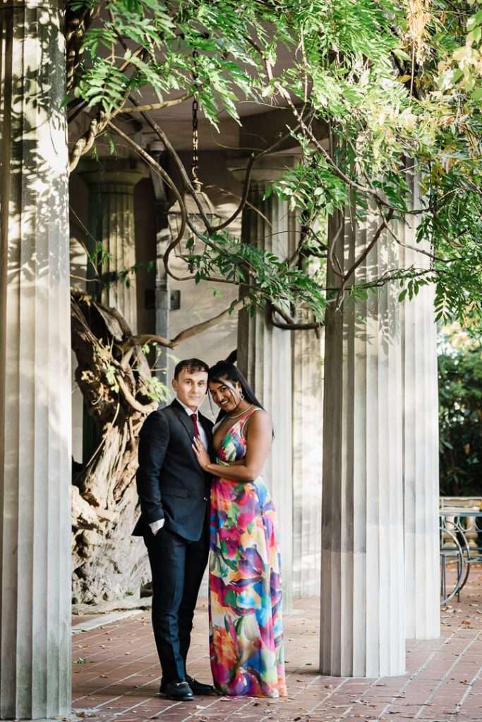 Romantic portrait of an engaged couple under the columned pergola at Van Vleck House, framed by twisting vines and golden afternoon light.