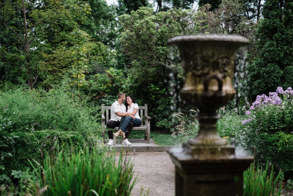 Engaged couple laughing on a bench past a stone urn at Van Vleck House Montclair NJ