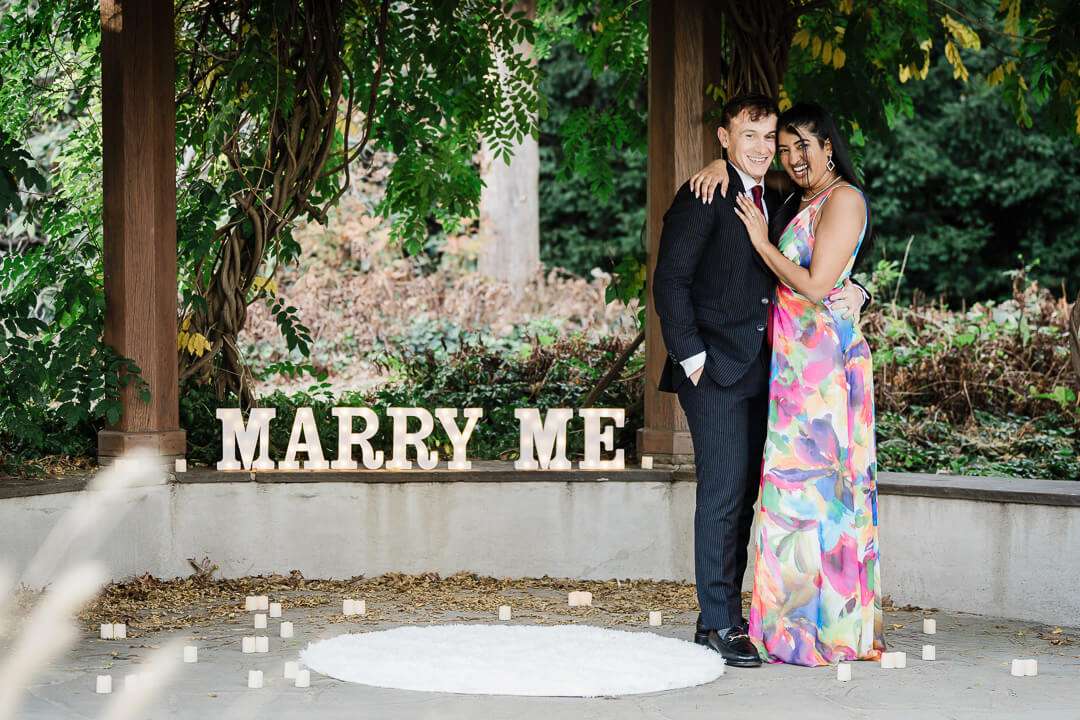 Couple standing under pergola at Van Vleck Gardens with “Marry Me” sign and candles surrounding them during surprise proposal
