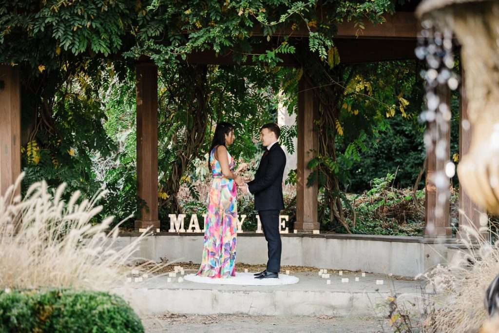 Couple standing face to face under the pergola at Van Vleck House moments before the proposal, surrounded by greenery and candles.