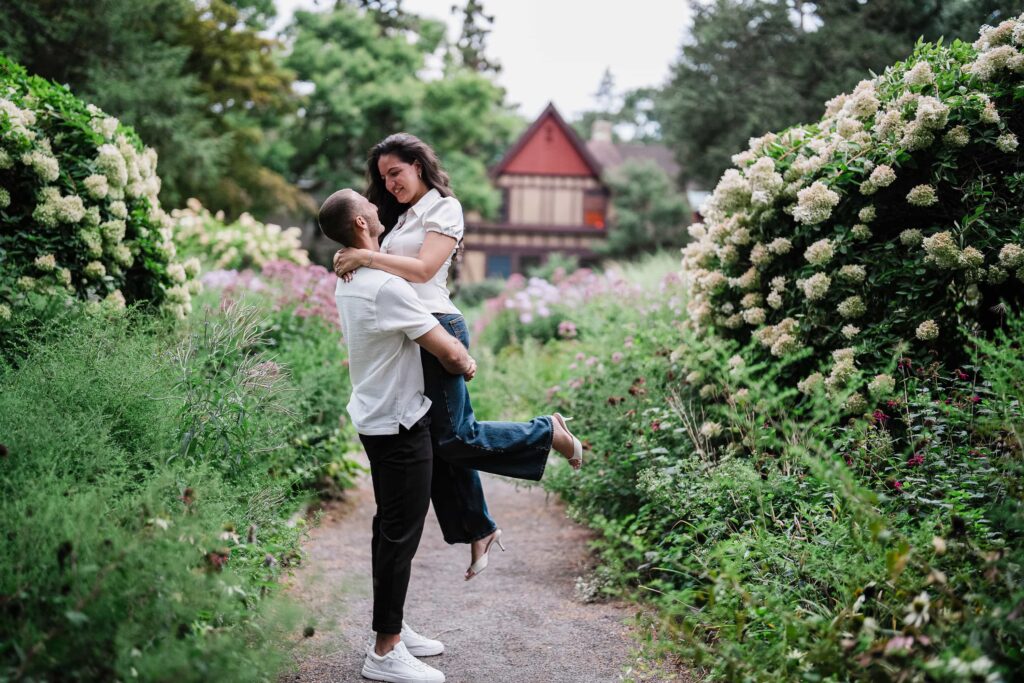 Man lifting his fiancee on a flower lined path at Van Vleck House Montclair NJ