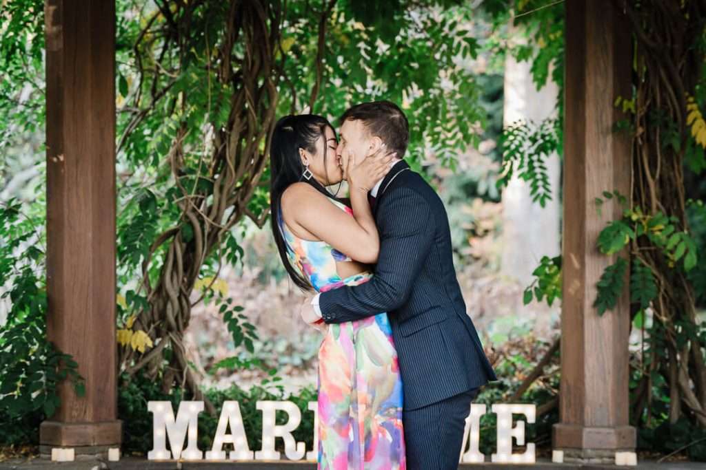Engaged couple kissing under the pergola at Van Vleck Gardens with vines and greenery surrounding them.