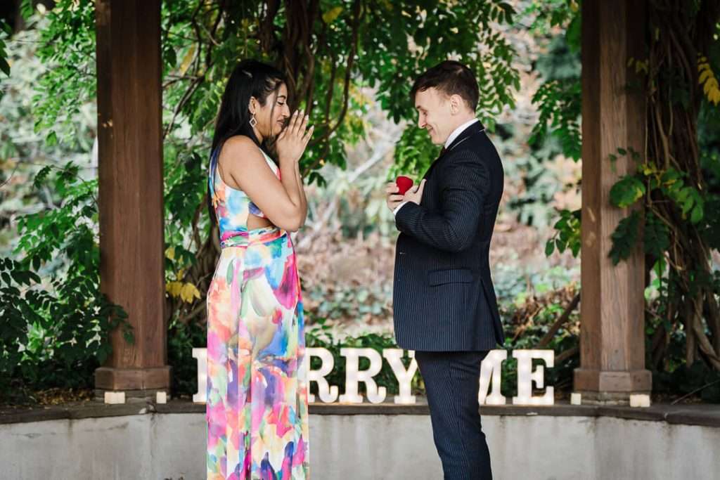 Groom-to-be holding a red ring box as his partner reacts with joyful surprise at Van Vleck House proposal.