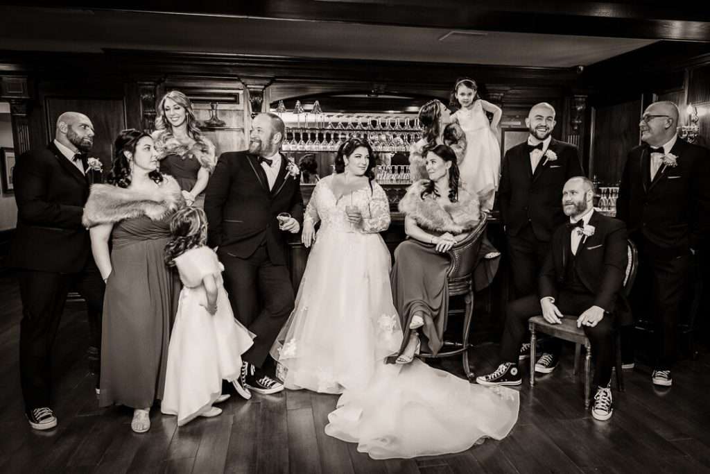 Bride and groom pose with their wedding party in a stylish bar lounge, wearing Converse sneakers and fur wraps in a fun and relaxed group portrait.