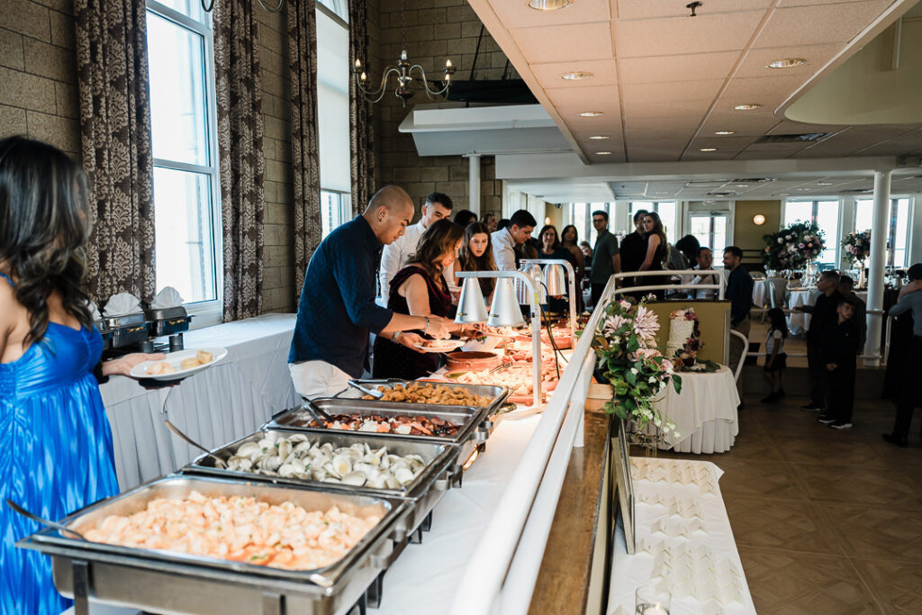 Guests serving themselves from a buffet featuring shrimp, clams, and assorted entrées during a waterfront engagement celebration at The Armory NJ Restaurant in Perth Amboy, photographed by Alex Kaplan Photography.
