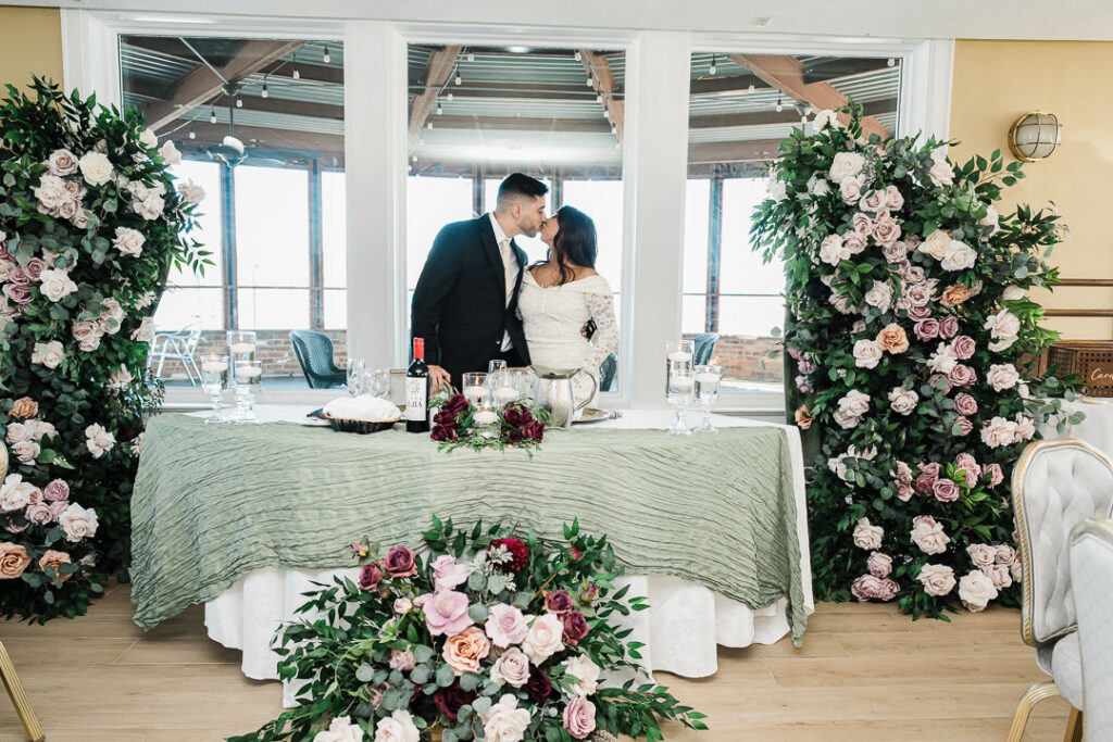 Bride-to-be Navi and fiancé Christian sharing a kiss by the waterfront windows surrounded by lush blush and mauve floral arrangements at The Armory NJ Restaurant in Perth Amboy, photographed by Alex Kaplan Photography.