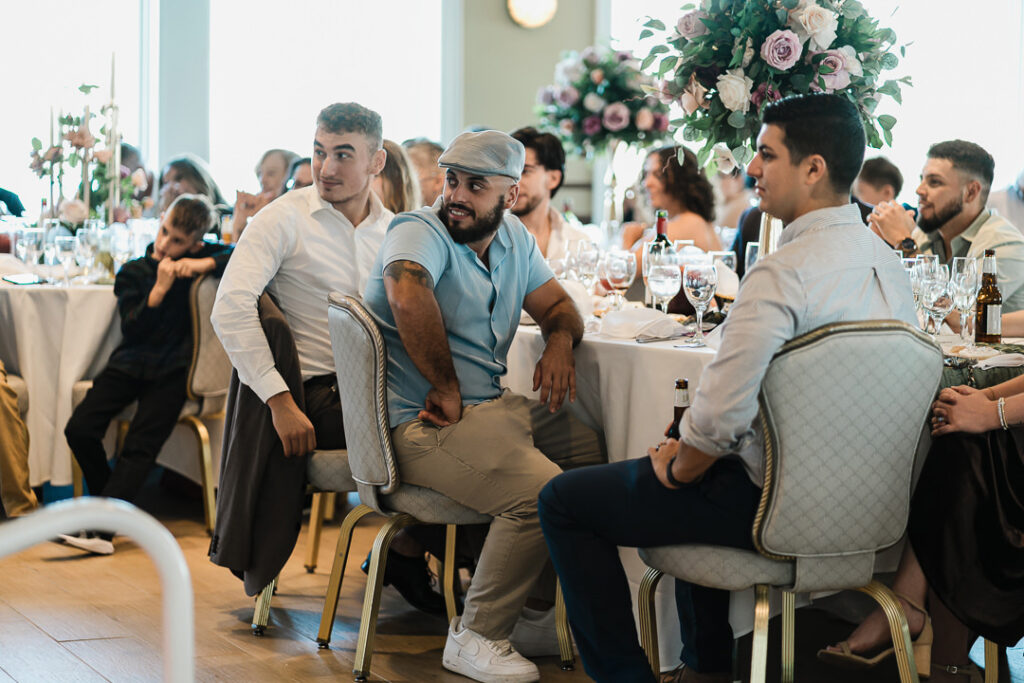 Guests seated at elegantly decorated tables with floral centerpieces enjoying heartfelt speeches and laughter during an engagement celebration at The Armory NJ Restaurant in Perth Amboy, captured by Alex Kaplan Photography.