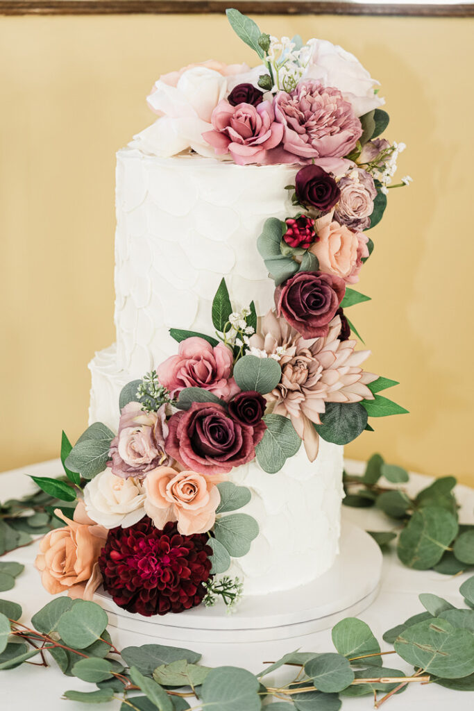 Three-tier white buttercream cake decorated with cascading fresh roses and dahlias in burgundy, blush, and peach tones with eucalyptus leaves, photographed by Alex Kaplan Photography at The Armory NJ Restaurant in Perth Amboy.