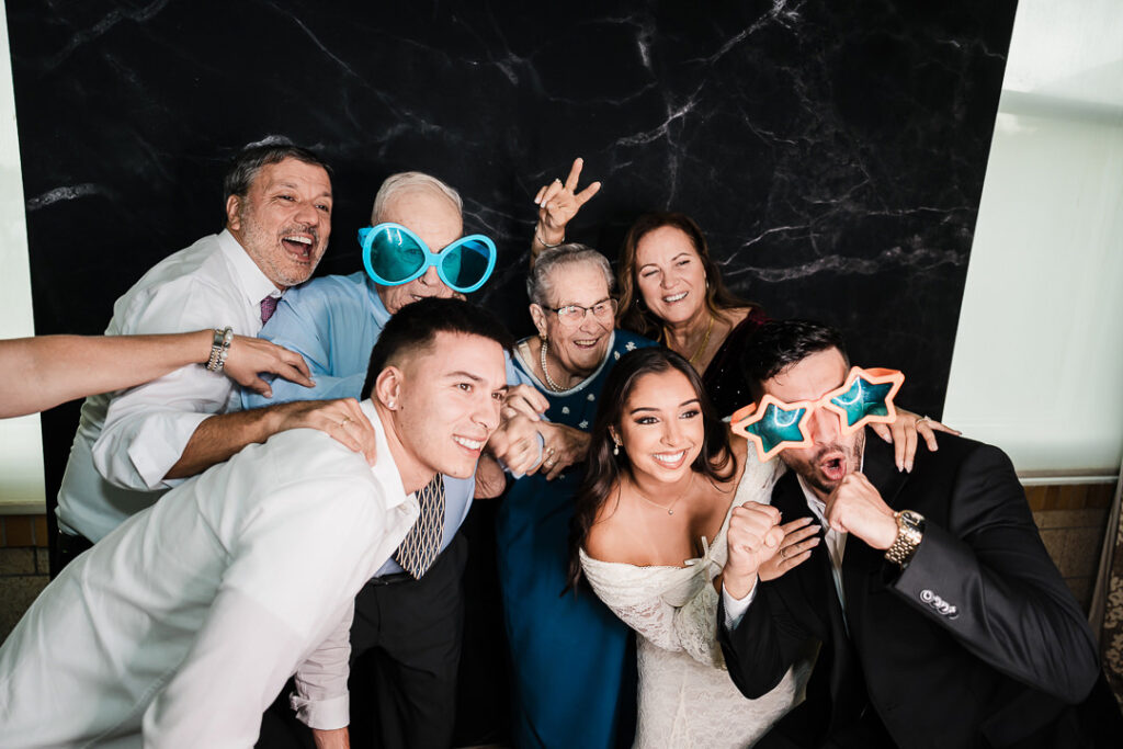 Bride and groom posing with family wearing fun photo booth props during wedding reception at The Armory NJ Restaurant in Perth Amboy, photographed by Alex Kaplan Photography.