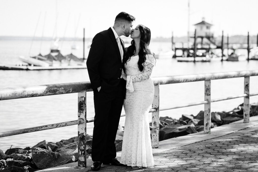 Black and white portrait of Navi and Christian kissing along the Perth Amboy marina railing with sailboats in the background, photographed by Alex Kaplan Photography during their engagement session at The Armory NJ Restaurant.