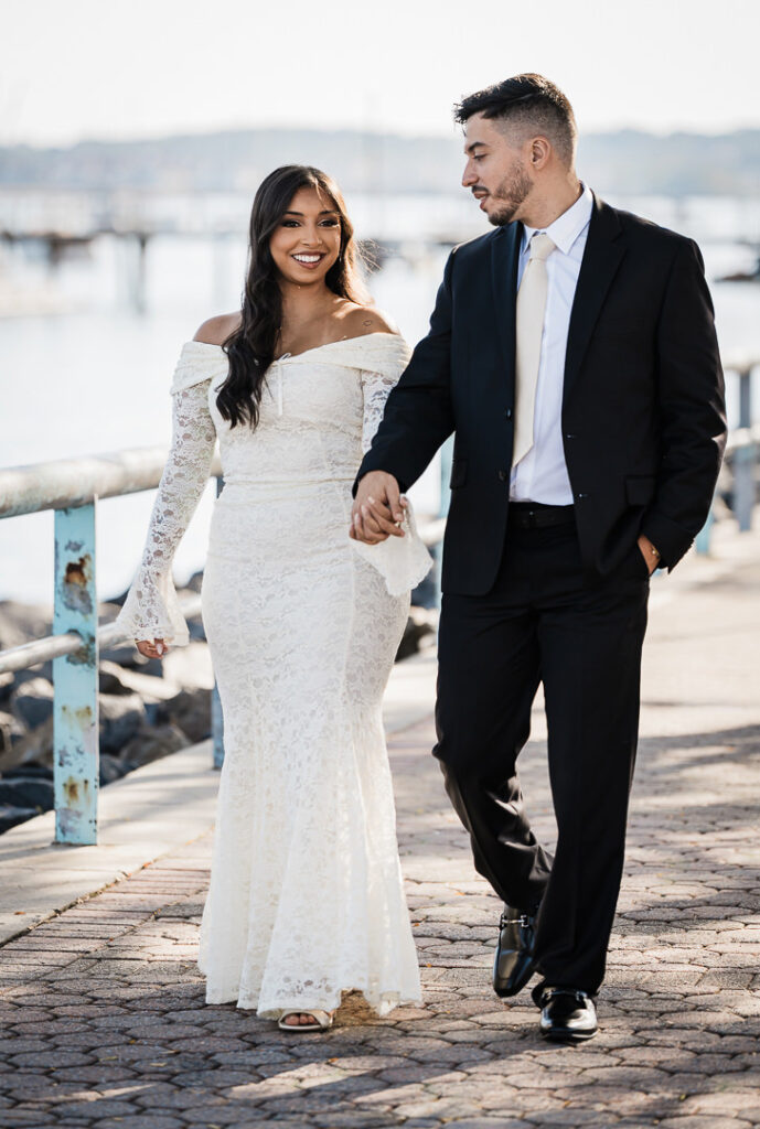 Engaged couple walking hand in hand along the Perth Amboy waterfront in elegant attire, featuring a white lace gown and black suit, captured by Alex Kaplan Photography at The Armory NJ Restaurant.