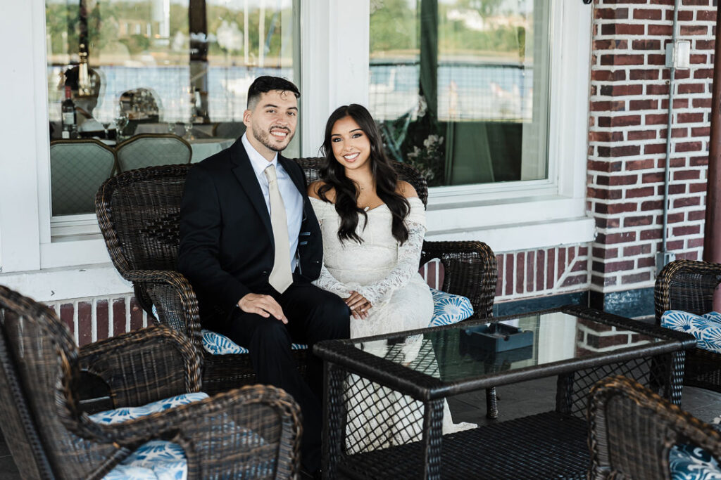 Engaged couple sitting together on the outdoor patio of The Armory NJ Restaurant in Perth Amboy, smiling and dressed elegantly for their waterfront celebration, photographed by Alex Kaplan Photography.