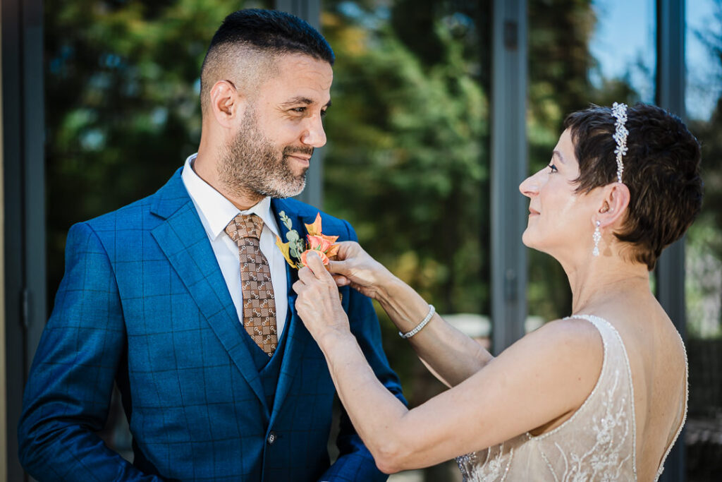 Bride pinning boutonniere on the groom’s suit outside Mohawk House in natural afternoon light.